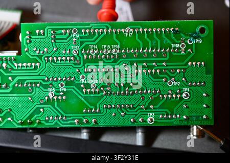 Technician Testing Circuit Board Tracks with Multimeter Probe Close Up. High quality photoMacro shot of the backside of a green printed circuit board (PCB) showing soldered joints, copper tracks, and marked test points (TP). A technician uses a probe tool to test connections and diagnose electrical faults. Perfect for concepts of electronics diagnostics, hardware repair, soldering quality inspection, and engineering. Stock Photo