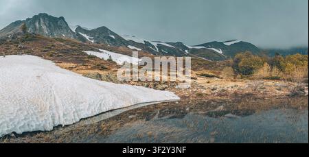 Mountain landscape showing a cold lake reflecting snowy peaks and dry grass, representing the transition from winter to spring Stock Photo