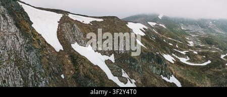 Panoramic view of a mountain range with patches of snow and green grass, covered in mist on a cloudy day Stock Photo