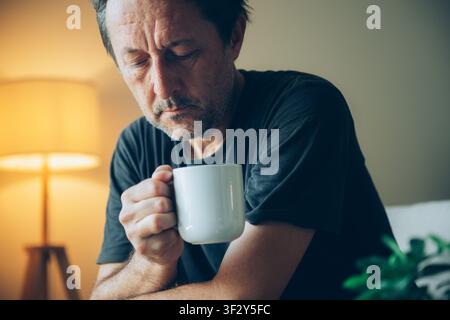 A contemplative middle-aged man drinking from a coffee mug in warm living room light, appearing thoughtful and alone as he quietly reflects during an Stock Photo