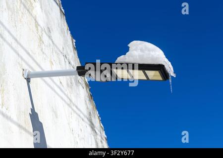 Architectural detail of a modern black LED street lamp mounted on a peeling white-painted wall, heavy fresh snow piled thickly on top, selective focus Stock Photo