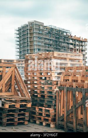 Stacks of wooden shipping pallets and crates at a construction site with a large building under development behind them. Selective focus. Stock Photo