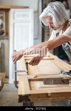 Talented man restorer in woodworking shop fixating antique wooden door at workbench using clamps Stock Photo