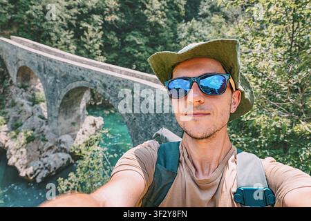 Young man wearing sunglasses and hat taking a selfie while exploring nature and hiking near an old stone bridge Stock Photo