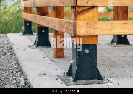 Close-up of timber fence posts mounted in black steel bases bolted to a concrete pad, with gravel edging and greenery behind. Stock Photo