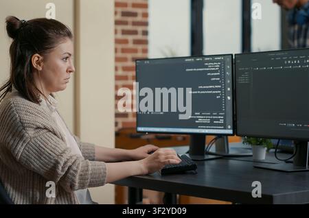 Focused programmer writes code while looking at multiple computer screens, with programming language algorithms and teamwork in the background. Stock Photo