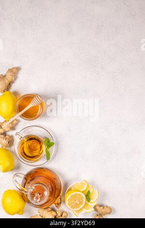 Hot ginger lemon tea brewing in a glass teapot and served in a cup alongside fresh lemons, sliced ginger root, a bowl of honey with dipper, and mint l Stock Photo