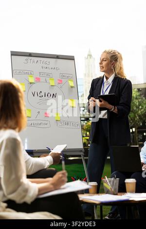 Businesswoman presenting strategy roadmap on vision board during informal meeting, pointing at whiteboard with sticky notes. Outdoor team workshop planning session. Stock Photo