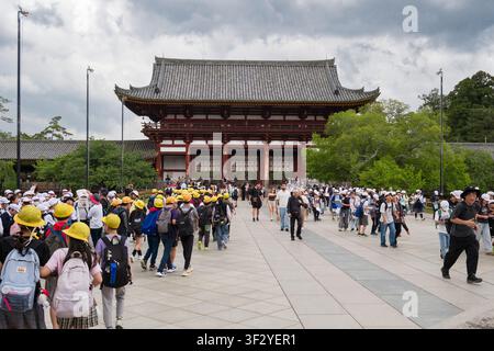 A large crowd of visitors, including many students wearing yellow safety helmets, gathers at the main gate of Tōdai-ji Buddhist temple in Nara, Japan. Stock Photo