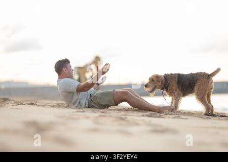 Male dog owner sitting on sandy beach interacts with his pet Airedale Terrier during sunset, with water and distant figures in the background Stock Photo