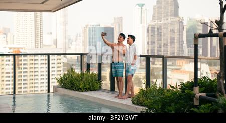 Gay couple taking selfie enjoying summer vacation at rooftop swimming pool Stock Photo