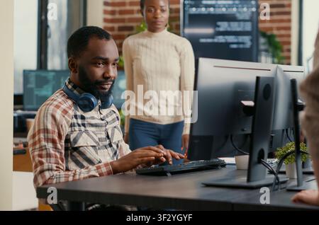 Smiling software developer wearing wireless headphones smiling working at computer screen while typing at a computer, showing a smiling developer workflow with code, headphones and IT office screens. Stock Photo