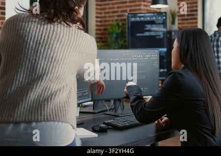Programmer pointing pencil at computer screen with software compiling code explaining source algorithm to coworker standing next to desk around computer screens, showing developer teamwork, source. Stock Photo