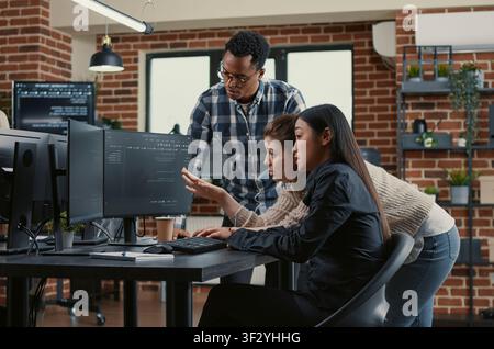 Mixed software engineers brainstorm cloud computing interface ideas while reviewing running code on computer screens in a development office. Stock Photo