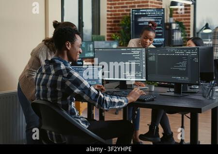 Software developer reviews running code on multiple screens, takes off his glasses and celebrates with a colleague high five during a successful coding moment. Stock Photo