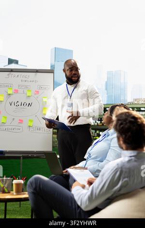 Black presenter explaining strategy roadmap with clipboard in hand, presenting ideas on sticky notes and whiteboard during collaborative planning discussion. Informal meeting. Stock Photo