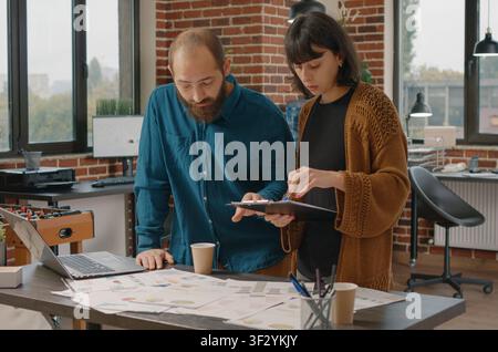 Business colleagues use paper charts to create a marketing strategy, with a woman checking clipboard files while a man brainstorms presentation ideas. Stock Photo