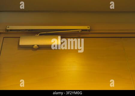 A silver mechanical automatic door closer mechanism mounted at the top of a closed wooden door inside an empty residential room. Stock Photo
