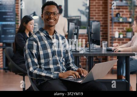 Portrait of artificial intelligence app developer sitting down typing on laptop fixing glasses looking up and smiling at camera, programer using portable computer innovating cloud computing. Stock Photo