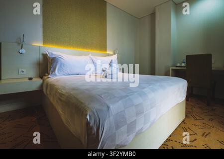 A neatly made double bed with white linens and a folded towel animal sitting empty inside a contemporary bedroom during the evening, featuring warm am Stock Photo