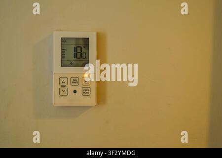 A white digital aircon remote control panel mounted on a plain wall shows a temperature reading of 18 degrees Celsius inside an empty residential room Stock Photo
