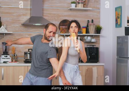 Carefree couple sings together in morning kitchen, linking breakfast, domestic routine and home comfort inside a domestic kitchen setting. Stock Photo