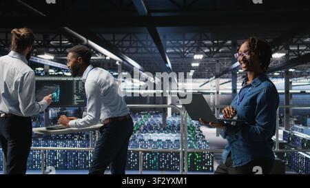 Happy woman in data center doing maintenance tasks on laptop, ensuring optimal performance. Engineering team reviewing diagnostics and applying patches to server room, improving stability, Stock Photo