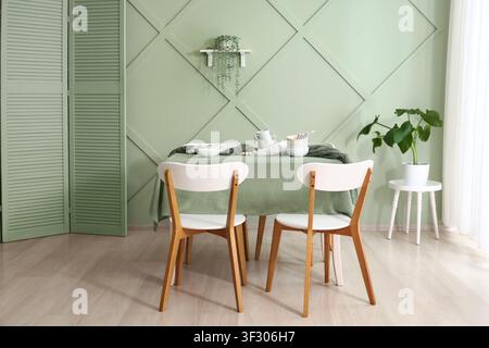 Dining room interior with modern wooden chairs and a table set with dishes against a sage green wall, the room features a decorative wall paneling, a Stock Photo