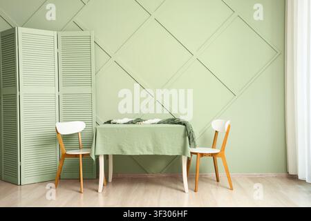 Minimalist dining room interior with green accent wall, wooden chairs, and a table covered with a tablecloth Stock Photo