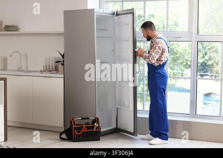 Professional repairman in blue overalls fixing a refrigerator in a modern kitchen with a toolbox on the floor, a toolbox filled with various tools res Stock Photo