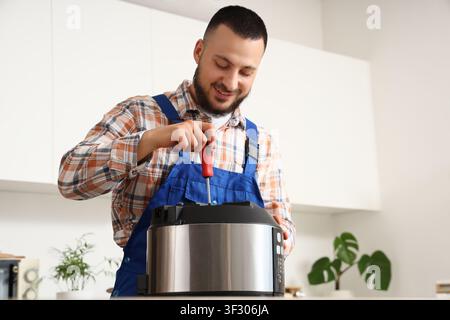 Smiling young repairman in blue overalls and plaid shirt fixing a kitchen appliance with a screwdriver, the professional handyman is focused on the ta Stock Photo