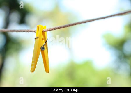 Single yellow plastic clothespin hanging on a brown rope clothesline with a blurred green and white background, the background is a soft blur of green Stock Photo