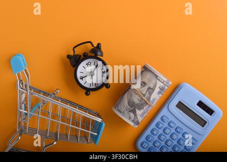 Shopping budget concept with mini cart, calculator, cash, and alarm clock on orange background, representing expense planning, financial control, time Stock Photo