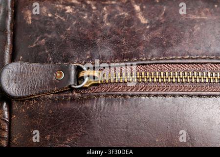 Macro shot of a brass zipper with metal teeth and leather pull tab on an aged dark brown leather bag featuring visible stitching and a textured patina Stock Photo