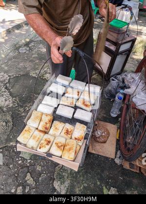 A street vendor using metal tongs to grill traditional Indonesian ketan bakar glutinous rice cakes on a wire mesh tray at an open-air market stall. Stock Photo