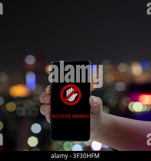 Hand holding a smartphone displaying an Access Denied message and stop icon against a blurred night city background. Ideal for cybersecurity, digital Stock Photo