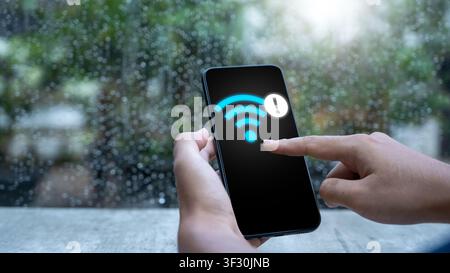 Close-up of hands holding a mobile phone showing a Wi-Fi error symbol against a rainy window background. Concept of bad internet connection and signal Stock Photo