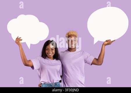 Millennial black couple holding empty speech bubbles on violet studio background, space for text Stock Photo