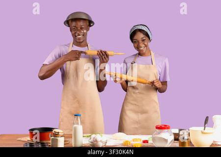 Positive black couple with rolling pins having fun while cooking, posing and looking at camera on violet background Stock Photo