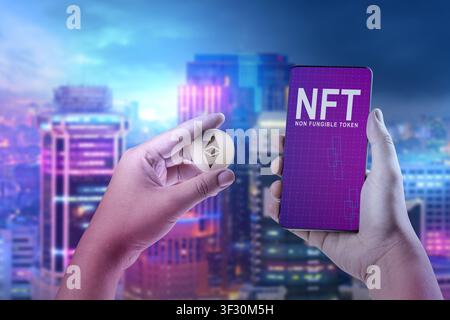 A person holding an Ethereum physical coin and a mobile phone displaying NFT text against a bokeh city night background. Concept of digital art and cr Stock Photo