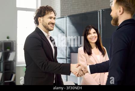 Two businessmen shake hands as smiling colleague watches in bright modern office Stock Photo
