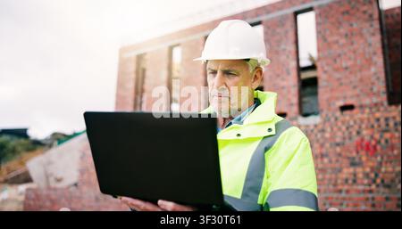 Reading, man and construction worker on site with laptop for research on building renovation. Computer, plan and mature civil engineer with feedback Stock Photo