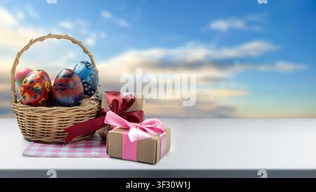 Festive composition featuring a wicker basket with hand-painted Easter eggs and wrapped gift boxes on a white tabletop against a blurred sky backgroun Stock Photo