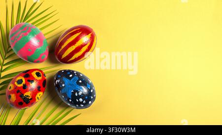 Collection of four uniquely hand-painted Easter eggs resting on green palm leaf. The scene features a vibrant yellow background with ample negative sp Stock Photo