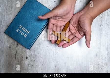 Top-down view of cupped hands holding a wooden cross next to a blue leather-bound Holy Bible on a grey textured background Stock Photo
