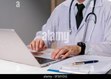 Medical practitioner with stethoscope and smartwatch working on laptop. Doctor consulting patient online or writing prescription. Modern hospital offi Stock Photo