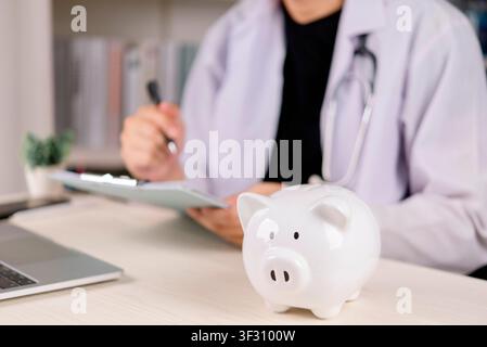 White piggy bank on desk with blurred doctor using stethoscope in background. Healthcare medical cost, health insurance savings, hospital clinic budge Stock Photo
