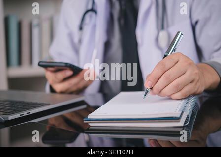 doctor hands holding smartphone and writing on notebook with stethoscope and laptop on desk. Concept of telemedicine, medical consultation, online app Stock Photo