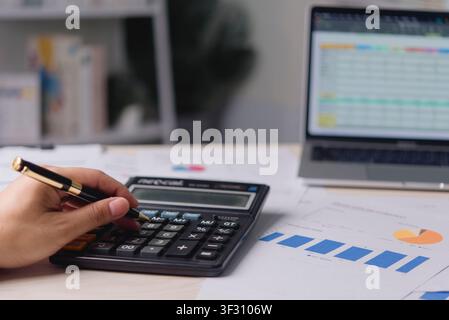 Close up accountant hand holding pen and using calculator for financial data analysis. Business workspace with laptop displaying spreadsheet and marke Stock Photo