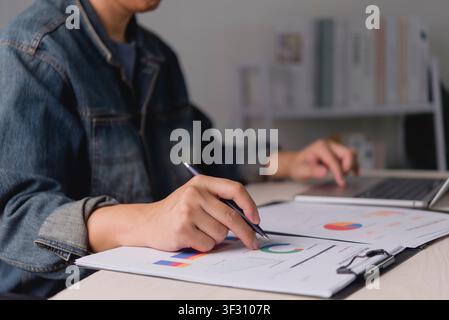 freelancer man hand holding pen pointing at business document paper, analyzing marketing chart and graph data while using laptop. Startup entrepreneur Stock Photo